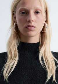 Schwarzer, strukturierten Pullover mit hohen Kragen; Modell mit langen blonden Haaren und großen silbernen Creolen. Neutrale Hintergrund. Minimale Details.