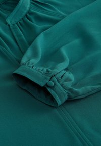 Îmbrăcăminte cu mâneci lungi dintr-un material teal închis, cu o textură moale. Manseta strânsă cu detaliu de nasture și pliere subtilă aproape de guler.