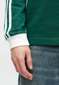 Zelená košeľa s dlhým rukávom a bielymi pruhmi na rukávoch, s bielymi manžetami. Ruka spočíva na svetlom denimovom materiáli.