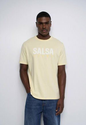 Jeune homme debout, les mains dans les poches, portant un t-shirt "SALSA JEANS" jaune clair et un jean bleu sur un fond blanc uni.