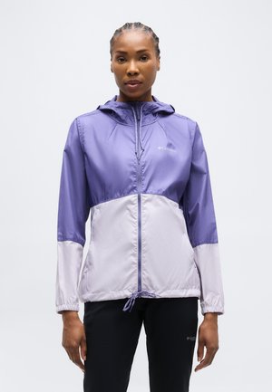 Donna in piedi che indossa una giacca con cappuccio viola e grigio chiaro e pantaloni neri, rivolta verso l'obiettivo su uno sfondo semplice.