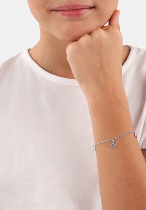 Silberne Charmarmband mit Kettendesign, das einen Schmetterling und einen kleinen roten Akzent zeigt. Am nackten Handgelenk vor einem hellen Stoffhintergrund getragen.