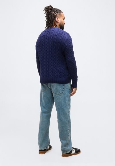Άνδρας με ντραντλoκς στέκεται με την πλάτη προς τα εμπρός φορώντας μπλε ναυτικό πουλόβερ με πλέξη καλωδίου, ανοιχτόχρωμα τζιν και μαύρα και λευκά αθλητικά παπούτσια.