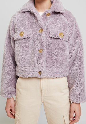 Lilla, fuzzy kort jakke med store brune knapper og klaplommer, båret med cremefarvede bukser.