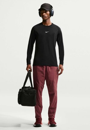 Mand iført sort langærmet skjorte, sort kasket, høretelefoner, vinrøde bukser og sneakers, der holder en sort duffeltaske i venstre hånd.