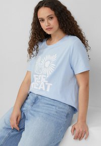 Lichtblauw katoenen T-shirt met korte mouwen, met een witte grafische print van bloemen en de tekst "FEEL GREAT." Gecombineerd met een lichte spijkerbroek.