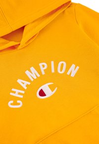 Felpa gialla con cappuccio realizzata in tessuto morbido. Presenta il testo e il logo "CHAMPION" ricamati in bianco, con una tasca frontale per lo stoccaggio.