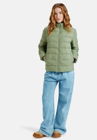 Grön vadderad pufferjacka med hög krage, full dragkedja och sidofickor, kombinerad med ljusblå vida jeans och bruna skor.