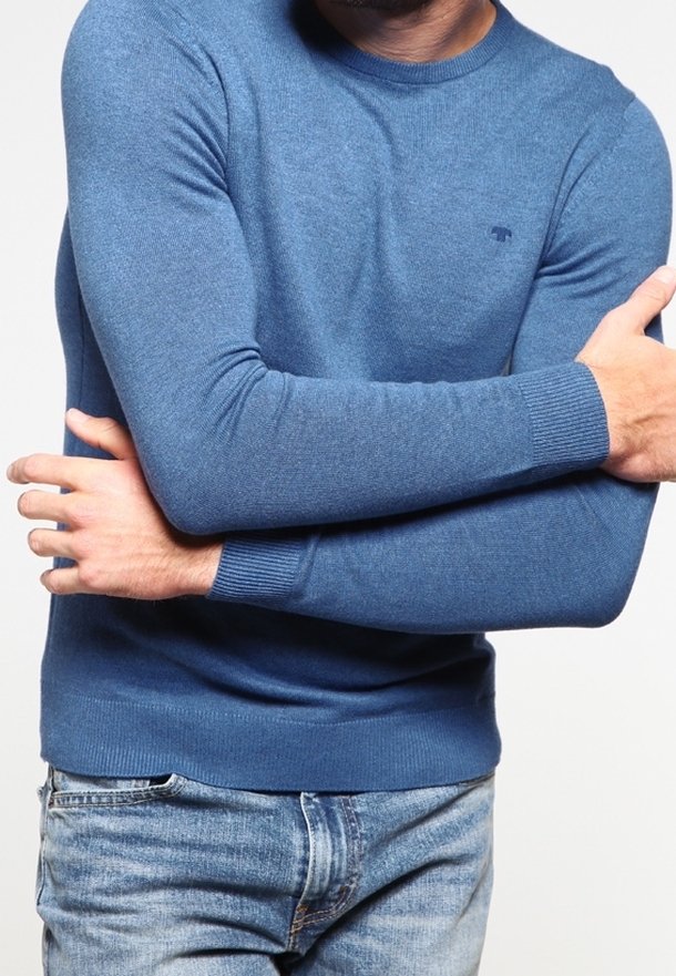 Pull bleu en tricot avec un col rond et des poignets côtelés, présentant un petit logo sur la poitrine. Associé à un jean en denim clair.