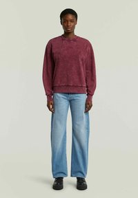 Felpa in cotone marrone con maniche raglan, colletto e orlo a costine, abbinata a jeans a gamba larga blu chiaro e scarpe nere.