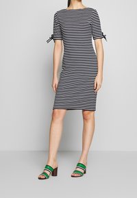 Vestido a rayas en azul marino y blanco, de manga corta, con lazos laterales, forma ajustada y dobladillo a la altura de la rodilla, combinado con sandalias de tacón bloque a rayas verdes y blancas.