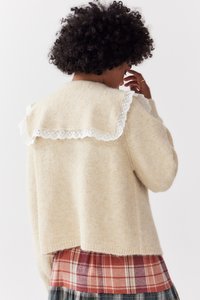 Vrouw met krullend haar in een beige gebreide trui met witte kantdetail en een rood-grijze geruite rok, met haar gezicht naar weg gekeerd en haar hand bij haar gezicht.