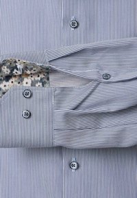 Blauw-wit gestreepte overhemden met bloemenmotief aan de binnenkant van de manchetten, een knoopkraag en een detail met twee knopen op de manchetten.