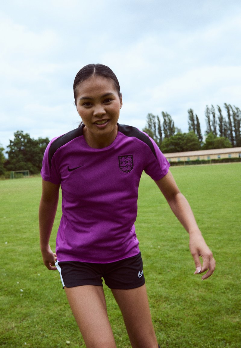 Purple Nike sports shirt with short sleeves, featuring a black accent on the shoulders and a crest; paired with black athletic shorts.