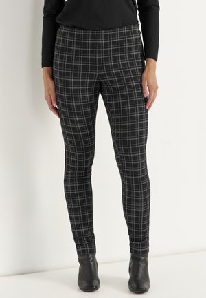 Persoon draagt zwarte en grijze geruite strakke broek gecombineerd met zwarte laarzen en een zwart top met lange mouwen, staand tegen een witte achtergrond.