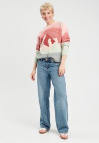 Strickpullover mit Bergdesign in Pastellfarben, kombiniert mit hellblauen weit geschnittenen Jeans und rosa Schlupfschuhen. Einfache Hintergrund.