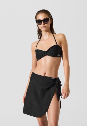 Čierny bikini top s prekrúteným dizajnom, doplnený čiernou sukňou voľného strihu. Modelka má tmavé slnečné okuliare a mokré vlasy.