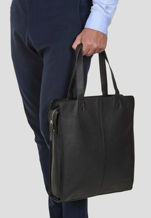 Black leather tote bag with textured surface, double handles, and a zip closure. Displayed being held with navy trousers.