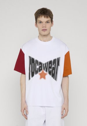 Nuori mies, joka käyttää valkoista oversize T-paitaa, jossa on punaiset ja oranssit hihat ja mustaa vääristynyttä tekstiä oranssin tähden kanssa edessä.