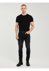 Homme debout portant un t-shirt noir ajusté, un jean noir déchiré avec une ceinture, et des bottes noires à lacets, sur un fond clair uni.