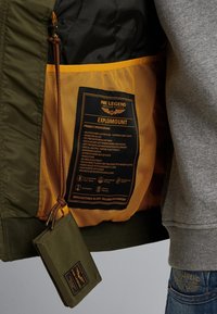 Veste de vol vert olive avec une doublure noire, dotée d'un intérieur en maille orange et d'une étiquette produit. Les détails du zip et des poches sont visibles.
