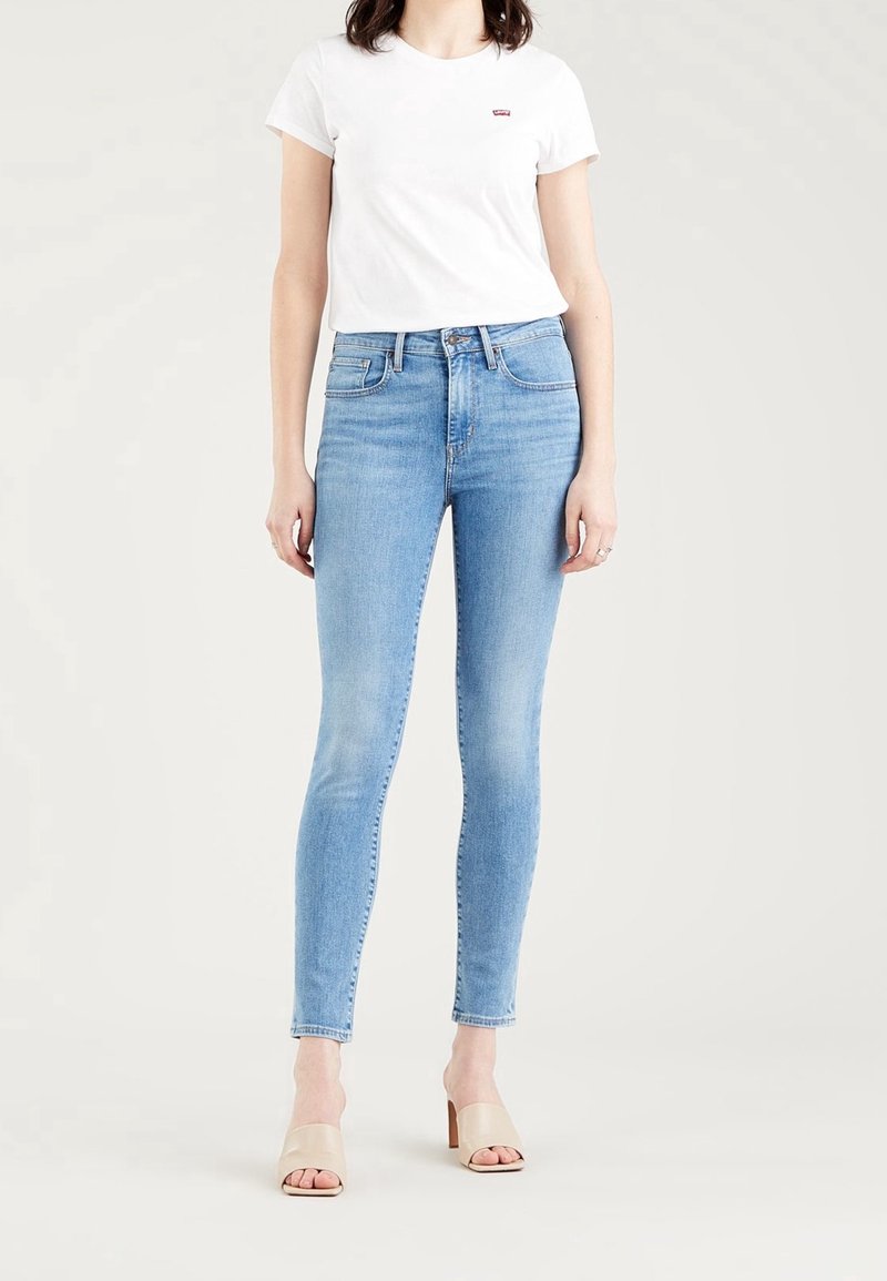 Femme portant un jean skinny bleu clair, un t-shirt blanc à manches courtes et des sandales à talons beiges, debout devant un fond clair uni.