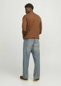 Brauner gestrickter Pullover mit Rundhalsausschnitt, zu weit geschnittenen hellblauen Jeans getragen. Braune Schuhe sichtbar, mit einem schlichten Hintergrund.