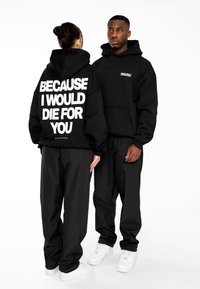 Sudaderas negras con gran texto blanco en la parte trasera y un pequeño logo en la parte delantera, combinadas con pantalones negros holgados y zapatillas blancas.