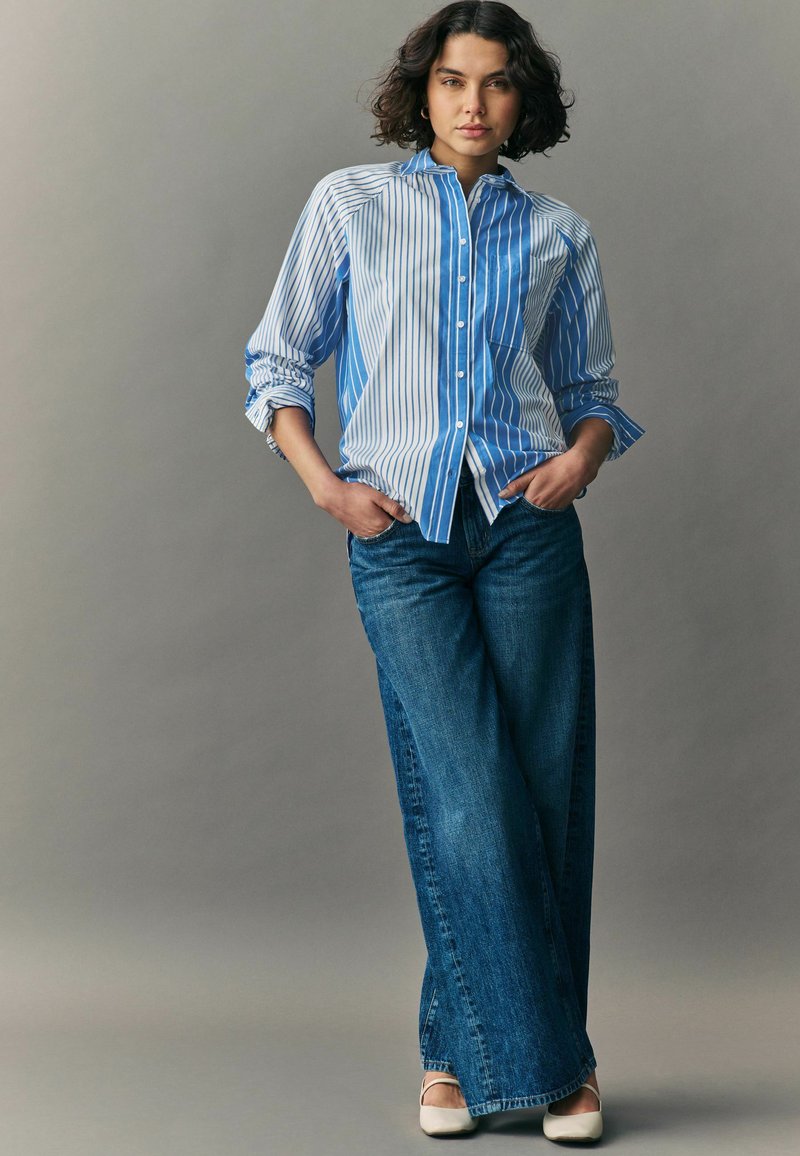 Camicia a righe blu e bianche con maniche arrotolate, abbinata a jeans blu a gamba larga e scarpe di colore chiaro.