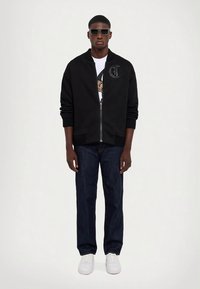 Svart bomberjacka med dragkedja, ribbade muddar, lättviktig tyg, med en logodetalj på bröstet, kombinerad med mörka jeans och vita sneakers.