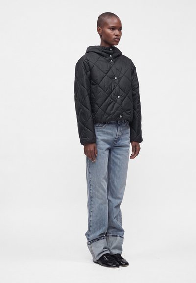 Personne portant une veste matelassée noire à capuche, un jean bleu clair à jambes larges avec revers, et des chaussures noires, debout devant un fond blanc uni.