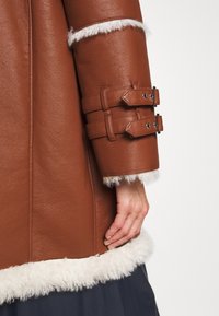 Rukáv hnědého koženého kabátu s bílým kožíškovým lemem a dvěma přezkami, zobrazený na paži a ruce osoby na neutrálním pozadí.