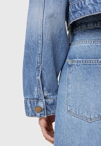 Chaqueta de mezclilla de color azul ligeramente desvaído, con bordes deshilachados y un detalle de botón en el puño. Costuras visibles que acentúan las costuras.