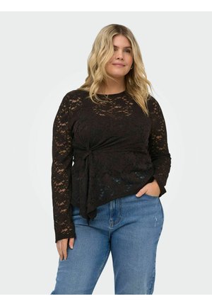 Vaaleahiuksinen nainen, jolla on päällä pitkähihainen musta pitsitoppi, jossa on solmudetalji, ja siniset farkut, seisoo käsi taskussa.