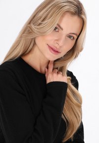 Pulover negru tricotat cu detalii texturate, purtat de o persoană cu păr lung, ondulat și blond, cu mâna odihnindu-se aproape de guler, fundal neutru.