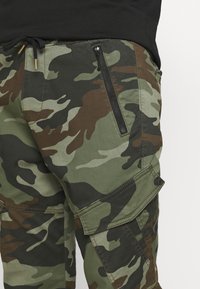 Vihreät ja ruskeat maastokuvioiset cargo-housut, joissa on vetoketjullinen tasku, säädettävä vyötärönauha ja sivutaskujen vahvistetut saumat.