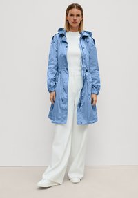 Giacca in nylon azzurro chiaro con vita regolabile, polsini elasticizzati e cappuccio, indossata sopra un top in maglia bianco e pantaloni larghi color crema.