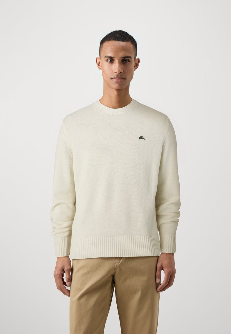 Kremeni pleteni džemper s rebrastim rubom i manžetama, sa malim zelenim Lacoste logotipom na prsima. Prilagođen kroj, klasični okrugli izrez.