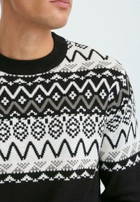 Gestrickter Pullover mit einem geometrischen Muster in Schwarz, Weiß und Grau. Rippbündchen am schwarzen Ausschnitt, mittleres Gewicht und kurze Ärmel.