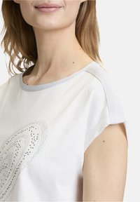 Weißes Baumwoll-T-Shirt mit kurzen Ärmeln, das silberne metallische Akzente und ein dekoratives Strassmuster auf der Vorderseite aufweist.