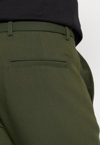 Olivgrüne Hosen aus strukturiertem Stoff, mit einer Gesäßtasche, die mit einem geraden Stich versehen ist, und einer Gürtelschlaufe.