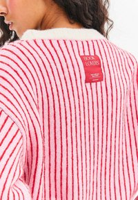 Proužkovaný pletený svetr v růžové a červené barvě. Má měkký, chlupatý bílý límec a na zadní straně červenou obdélníkovou nášivku s nápisem "BOOK LOVERS".