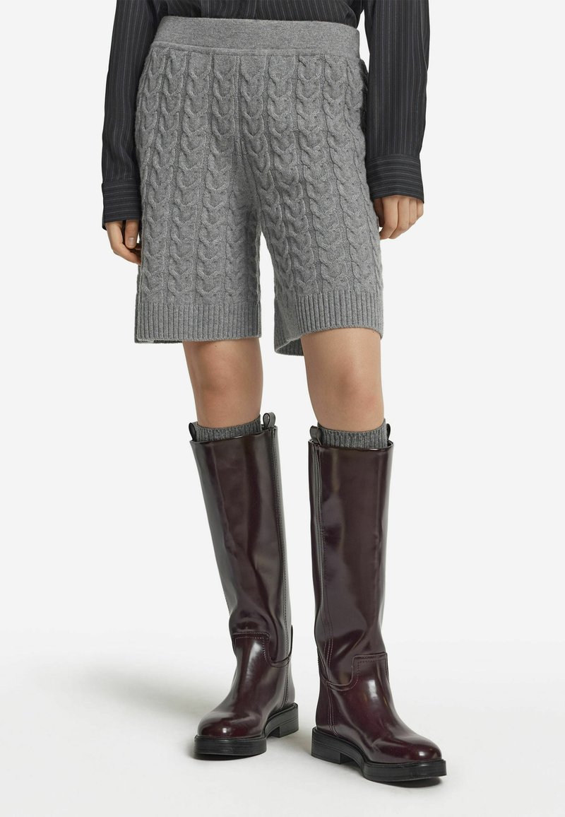 Graue, kabelgestrickte Shorts mit einem gerippten Bund, kombiniert mit knielangen burgunderfarbenen Stiefeln mit glänzender Oberfläche und flacher Sohle.