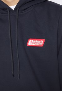 Sudadera azul marino con cordones negros y un parche rojo y blanco que dice "Carhartt Services" en el pecho.