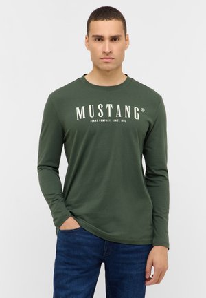 Grünes Langarm-T-Shirt mit dem Aufdruck "MUSTANG" in weißer Schrift. Hat einen Rundhalsausschnitt und eine lockere Passform. Kombination mit blauen Jeans.