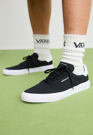Jambes portant des chaussettes blanches Vans et des baskets noires Adidas avec des semelles blanches, debout sur un sol vert.