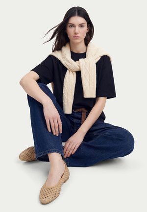 Donna con capelli lunghi e scuri seduta a gambe incrociate, con indosso una t-shirt nera, jeans blu, ballerine beige intrecciate e un maglione color crema drappeggiato sulle spalle.