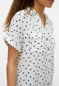 Chemise blanche à manches courtes avec des pois navy, dotée d'un bouton sur le devant, de deux poches poitrine et d'une coupe décontractée.