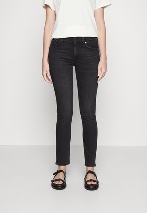 Vrouw draagt zwarte skinny jeans, wit topje met korte mouwen en zwarte sandalen, staand tegen een effen lichte achtergrond.