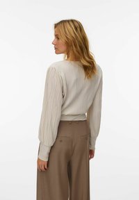 Lichtbeige blouse met verticale strepen, lange pofmouwen en een getailleerde taille. Gecombineerd met hoge, bruine broeken met twee achterzakken.